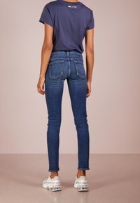 Jeans ajustados de color azul con una cintura a media altura y un lavado oscuro. Llevados con una camiseta azul marino y zapatillas blancas con suelas transparentes y texturizadas.