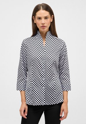 Kvinde iført sort og hvid skjorte med geometrisk mønster og trekvartærmer, stående foran en ensfarvet baggrund.