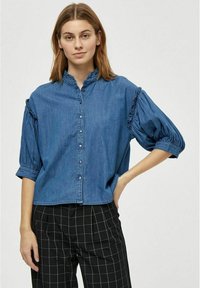 Denim bluzka z visokim ovratnikom, z gumbi spredaj in kratkimi napihnjenimi rokavi, ki imajo nagubane detajle. Nosena z črno-belimi karirastimi hlačami.