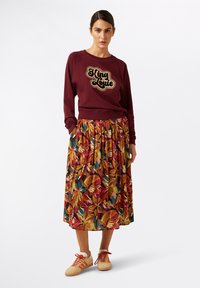 Bordeaux sweatshirt met "King Louie" opdruk, gecombineerd met een gemusterd maxi-jurk met levendige bloemenontwerpen en zakken. Casual sneakers.