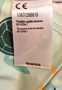 Hellblauem Stoff mit Mustern von Schildkröten und orangefarbenen Fischen; enthält ein Pflegeetikett mit CE-Kennzeichnung, Konformitätsdaten und Brandschutzwarnung.