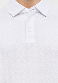 Λευκό γιακά πόλο και κουμπιά με διακριτικό υφασμάτινο μοτίβο ρόμβου και το γράμμα Α.