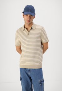 Beige pruhované polo tričko s krátkými rukávy, límcem a třemi knoflíky; spárováno s modrými cargo kalhotami a modrou kšiltovkou. Malé logo na hrudi.