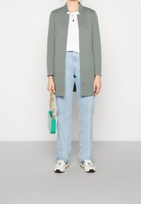 Cardigan gris ouvert sur le devant, t-shirt blanc, jean bleu clair, baskets blanches avec des accents verts, tenant un petit sac à main turquoise avec une chaîne.