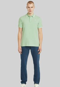 Ljusgrön bomullspolo med vit kant vid kragen och ärmarna, kombinerat med mörkblå jeans och vita sneakers.