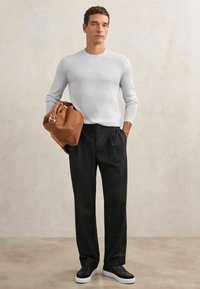 Jasnoszary obcisły sweter z długim rękawem, czarne plisowane spodnie, trzyma brązową zamszową torebkę i nosi czarne trampki z białymi podeszwami.