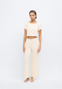 Σετ πιτζάμες δύο τεμαχίων σε ανοιχτό κρεμ χρώμα, με ένα κοντό τοπ με κυματιστά τελειώματα και σχέδια ροζ καρδιών, σε συνδυασμό με παντελόνι φαρδύ στο κάτω μέρος.