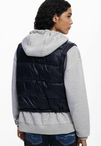 Mujer con cabello oscuro y rizado, vistiendo un chaleco acolchado negro sobre una sudadera gris y jeans azules, de perfil con una expresión neutral.