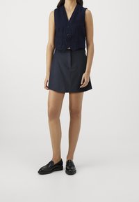 Gilet navy corto con scollo a V, tre bottoni e taschino sul petto, abbinato a una gonna A-line navy e mocassini neri. Tessuto liscio e vestibilità sartoriale.