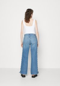 Vaqueros anchos azul con cintura alta, hechos de mezclilla. Presentan detalle de cordones en el lateral y dos bolsillos traseros. Llevados con una camiseta de tirantes blanca.