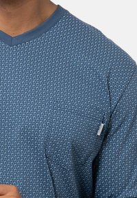 Sininen kuvioitu paita, jossa on timantin muotoinen kuvio, lyhyet hihat, V-pääntie ja rintatasku, jossa on etiketti. Teksturoitu puuvillakangas.