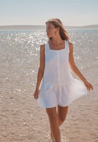 Biele šaty bez rukávov so štvorcovým výstrihom, prednými gombíkmi a volánovou spodnou časťou, oblečené na postave pri brehu vody.