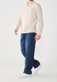 Beige linnen overhemd met knopen, blauwe denim jeans en witte sneakers. Het overhemd heeft een mandarijnkraag en lange mouwen. De jeans hebben een relaxte pasvorm.