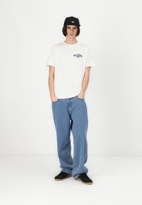 Hvit bomulls T-skjorte med en blå logo, kombinert med lyseblå baggy jeans. Svart caps er båret, og svarte sneakers med gummisåler.