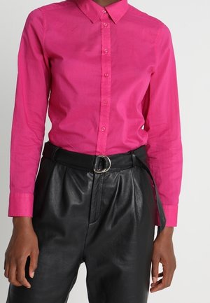 Kirkkaan pinkki pitkähihaisen napillisen paidan paita, joka on tökätty mustien korkeavyötäröisten nahkahousujen sisään. Vyössä on hopeinen kaksoisrenkaan solki.