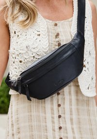 Černá kožená crossbody taška s elegantním designem, vybavená zipovým uzávěrem a texturovaným povrchem, nošena přes světlé pruhované šaty.