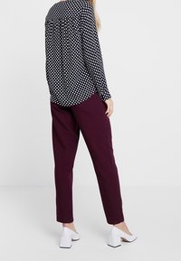 Blusa nera a pois con una vestibilità comoda, abbinata a pantaloni bordeaux a gamba affusolata e scarpe bianche con tacco a blocco. Vista da dietro.