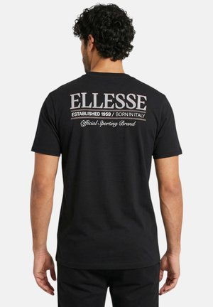 Homme aux cheveux bouclés portant un t-shirt noir avec le texte « ELLESSE Established 1959 / Born in Italy Official Sporting Brand » dans le dos.