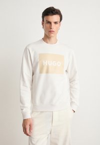 Baumwoll-Sweatshirt in hellbeige mit einem großen beigen rechteckigen Logo, auf dem "HUGO" in Weiß steht. Gerippter Rundhalsausschnitt und Bündchen.