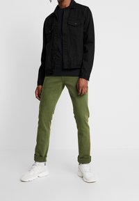 Svart denimjacka över en svart skjorta, tillsammans med rullade olivgröna byxor och vita sneakers. Klädseln har en avslappnad passform.