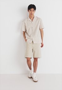 Camisa de manga corta a rayas en beige y blanco, combinada con pantalones cortos beige y zapatillas blancas. Diseño sencillo con un corte relajado y cierre de botones.