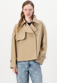 Giacca impermeabile beige tagliata con colletto oversize, chiusure a bottone e maniche larghe. Indossata sopra un top blu navy e jeans in denim con un motivo sbiadito.