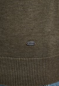 Maglione verde lavorato a maglia con orlo a coste, dotato di un piccolo logo in metallo nero. Texture morbida e spessore moderato.