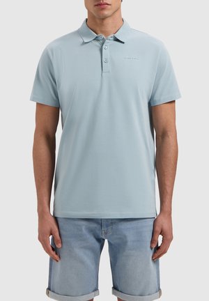 Lichtblauwe polo gemaakt van zachte stof, met een kraag, een sluiting met twee knoppen en subtiele branding op de borst. G gedragen met denim shorts.