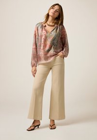 Bluza imprimată cu mânecă lungă în tonuri de roz și albastru, cu decolteu în V, asortată cu pantaloni bej cu picioare largi și sandale negre cu toc.