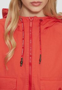 Rotkapuzenjacke mit durchgehendem Reißverschluss, verstellbaren Kordelzügen mit schwarzen Spitzen und zwei vorderen Taschen. Der Stoff wirkt glatt und leicht.