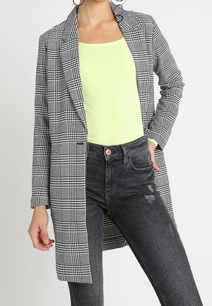 Femme portant un long blazer à carreaux noir et blanc, un haut vert néon, un jean noir taille haute, et de grandes boucles d'oreilles, debout.