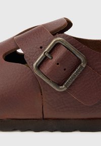 Sandale din piele maro cu bretea reglabilă și cataramă metalică. Prezintă un design decupat și o suprafață texturată. Talpa din cauciuc are un model în zigzag.