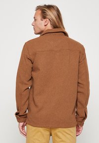 Brown textured shirt with a collar, button cuffs, and a straight hem. Herringbone pattern visible, featuring two back pockets.