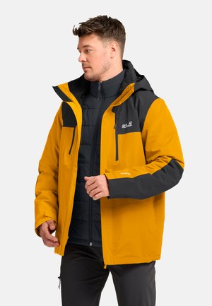 Mann trägt eine gelb-schwarze Outdoor-Jacke mit Kapuze und schwarze Hose, steht vor einem schlichten hellen Hintergrund.