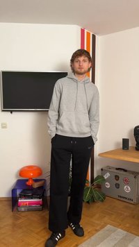 Szürke kapucnis pulóver kenguruzsebből, laza fekete nadrág és fekete sportcipő fehér csíkokkal. Egy színes akcentusokkal teli szobában áll.