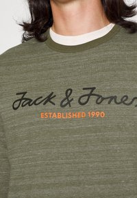 Felpa verde oliva con tessuto testurizzato, caratterizzata da testo in corsivo nero "Jack & Jones" e logo arancione "ESTABLISHED 1990".