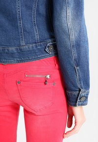 Tmavě modrá džínová bunda se zapínáním na knoflíky. Červené kalhoty s zipem na zadní kapse a malým ozdobným přívěskem. Hladká textura, přiléhavý střih.