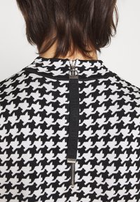 Vêtement en maille à motif pied-de-poule noir et blanc avec un col montant et une fermeture éclair dans le dos, comportant une tirette de marque texturée noire.