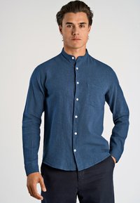 Camicetta blu a maniche lunghe con colletto alla cinese, tessuto testurizzato, taschino frontale e bottoni bianchi. Indossata con pantaloni scuri.