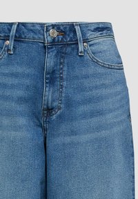 Niebieskie dżinsy z denimu, pokazujące pas, guzik, szlufki na pasek, przednie kieszenie oraz widoczne detale przeszyć.