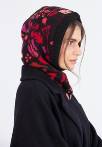 Vrouw in profiel draagt een zwarte jas en een zwarte capuchon met rode en roze bloemenpatronen, haarstrengen losjes bij haar gezicht, tegen een witte achtergrond.