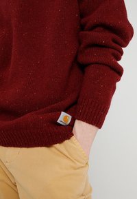 Burgundin neulepaita käännetyillä hihoilla, jossa on värikkäitä pisaroita ympäriinsä ja näkyvä Carhartt-logotagi helmassa, yhdessä khaki-housujen kanssa.