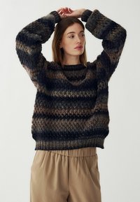 Pletený sveter v tmavých odtieňoch s textúrovaným vzorom v tvare šípok. Má okrúhly výstrih a široké rukávy, kombinovaný s béžovými nohavicami.