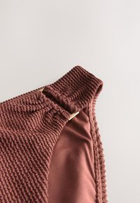 Gros plan d'une bretelle de maillot de bain en tissu rose profond texturé avec un élastique froncé et une boucle rectangulaire dorée sur un fond neutre.