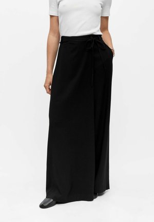 Femme portant un haut blanc à manches courtes et une longue jupe portefeuille noire avec une ceinture nouée et des chaussures noires sur fond blanc.