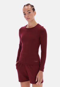 Top sportiv cu mânecă lungă, de culoare burgundă, fabricat dintr-un material moale și elastic, cu decolteu rotund și un detaliu subtil cu logo aproape de tiv.