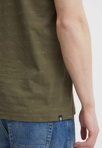 Olivgrünes Baumwoll-T-Shirt mit strukturierter Oberfläche und dezenten roten und weißen Sprenkeln. Mit einem Logo-Tag am Saum. Kombiniert mit blauer Jeans.