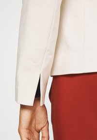 La manche d'un blazer blanc avec un détail de couture fendue, associé à un vêtement ajusté de couleur rouille. Le tissu semble lisse avec une coupe structurée.