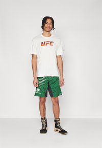 Fehér póló piros "UFC" grafikával, zöld mintás nadrág szürke részletekkel, és fekete csizmák arany részletekkel. Sportos szabás, laza stílus.