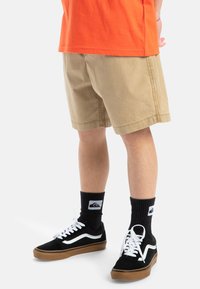 Pantalones cortos caqui con cinturilla elástica y bolsillos laterales, combinados con zapatillas negras con acentos blancos y calcetines de canalé negros.